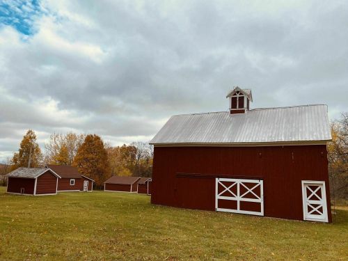 A red barn with a white roof stands in a grassy field, surrounded by smaller red buildings and autumn trees under a cloudy sky.