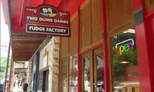 Sign for "Two Dumb Dames Fudge Factory" with an "open" neon sign in the window, set in a colorful storefront.