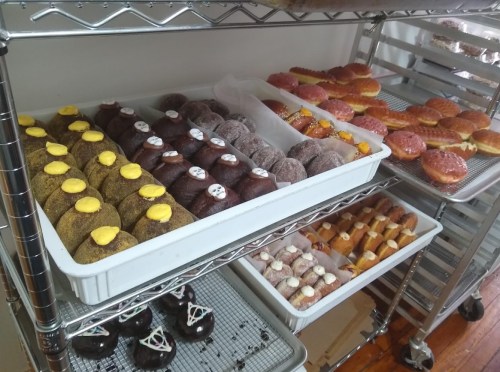 A variety of colorful donuts displayed on trays, including frosted and filled options, in a bakery setting.
