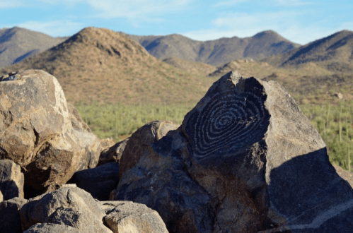 A close-up of a rock with a spiral petroglyph, set against a backdrop of mountains and desert vegetation.