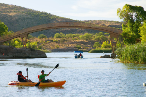 Two people kayak on a calm river under a wooden bridge, with a boat in the background and lush greenery along the banks.
