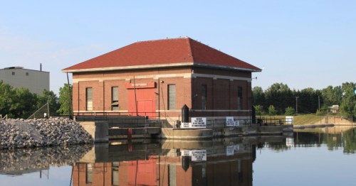 A historic brick powerhouse building with a red roof reflected in the still river water in Michigan.