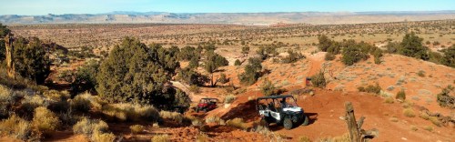 Off-road vehicles navigate a rugged, desert landscape with distant mountains and sparse vegetation under a clear blue sky.
