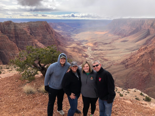 A group of four people poses at the edge of a canyon, with dramatic rock formations and a cloudy sky in the background.