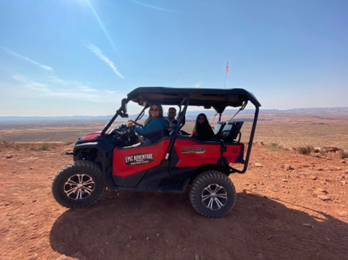 A group of three people in a red off-road vehicle on a rocky landscape with a clear blue sky in the background.