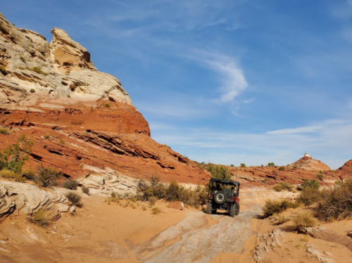 A jeep drives along a sandy path surrounded by red rock formations and blue skies.