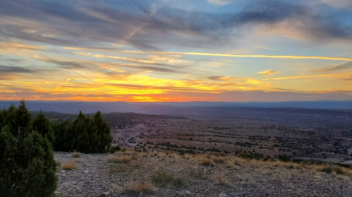 A vibrant sunset over a vast landscape, with rolling hills and scattered trees silhouetted against the colorful sky.