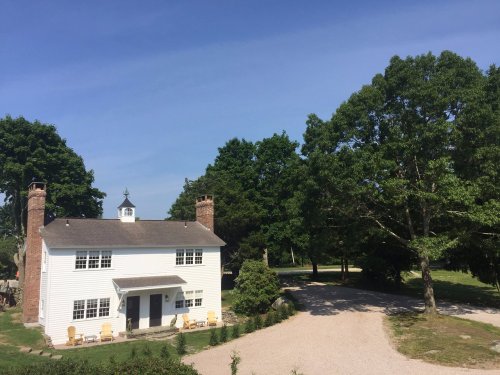 A white house with a cupola, surrounded by trees and a gravel driveway under a clear blue sky.