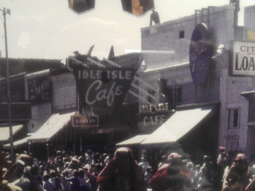 A vintage street scene featuring the Idle Isle Café and a crowd of people in front of various shops.