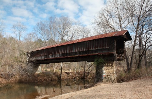 A rustic covered bridge with a rusty roof spans a calm river, surrounded by bare trees and a cloudy sky.
