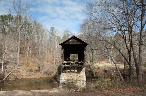 A rustic covered bridge stands over a calm river, surrounded by bare trees and a cloudy sky.