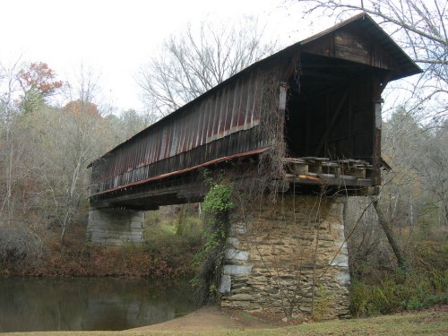 A weathered covered bridge stands over a calm river, surrounded by trees and autumn foliage.