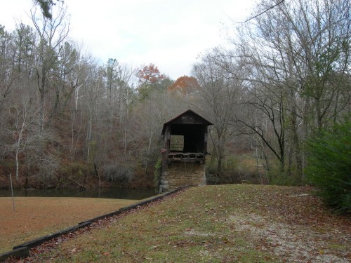 A rustic covered bridge stands over a calm river, surrounded by bare trees and autumn foliage.