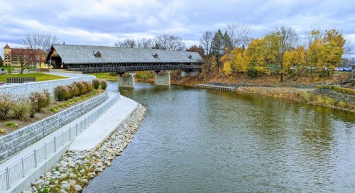A covered bridge spans a calm river, surrounded by autumn trees and a landscaped pathway.