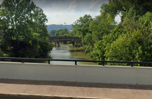 A serene river scene with a stone bridge in the background, surrounded by lush green trees and a clear sky.