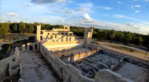 Aerial view of ancient ruins surrounded by greenery under a blue sky with scattered clouds.