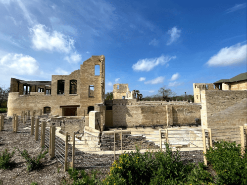 Ruins of a stone building under a blue sky, surrounded by greenery and a wooden fence.