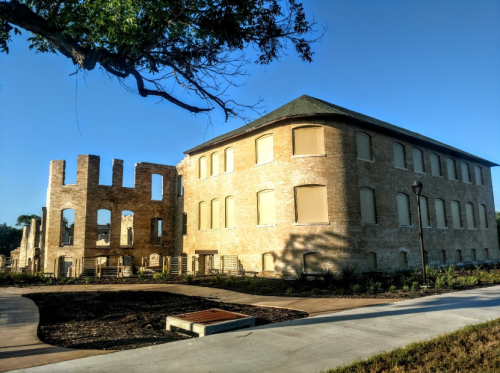 Historic stone building with large windows, partially ruined structure, and clear blue sky in the background.