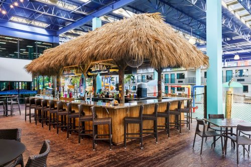 A tropical-themed bar with a thatched roof and seating, set in a vibrant indoor space with colorful decor.