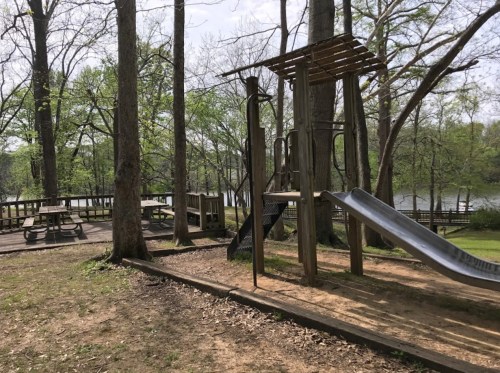 A playground with a slide and climbing structure, surrounded by trees and a picnic area near a lake.