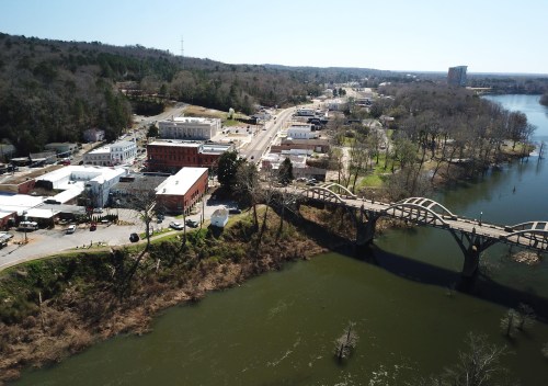 Aerial view of a river, bridge, and a small town with buildings and trees along the shoreline on a clear day.
