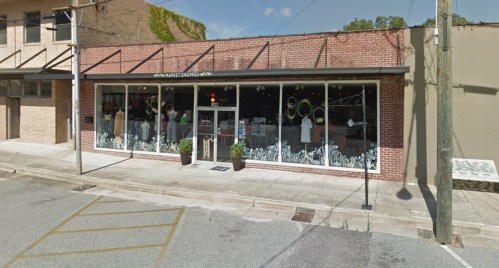 A storefront with large windows displaying clothing and decorations, featuring a brick facade and potted plants outside.