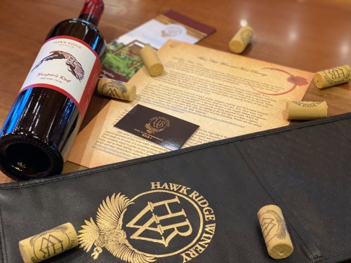 A bottle of Hawk Ridge Winery wine, corks, a tasting menu, and a branded bag on a wooden table.