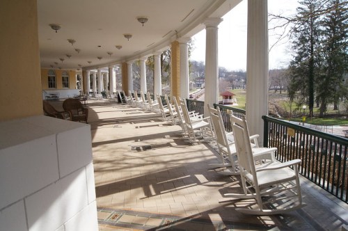 A row of white rocking chairs on a covered porch, with sunlight casting shadows on the tiled floor.