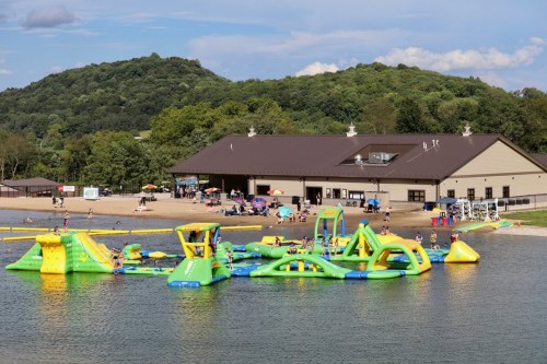 A colorful inflatable water park with people enjoying activities on a lake, surrounded by green hills and a building nearby.