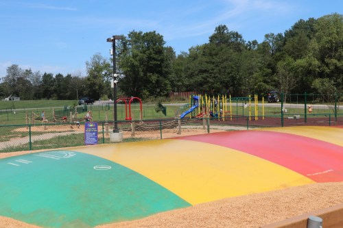 A colorful inflatable bounce pad in a playground with slides and swings in the background, surrounded by green trees.