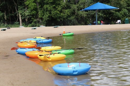 Colorful kayaks lined up on a sandy beach by a calm lake, with a blue canopy in the background.