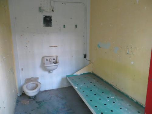 Empty, dimly lit room with a bare metal bed, a toilet, and peeling yellow paint on the walls.