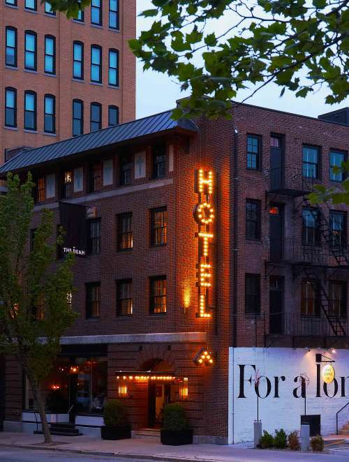 A brick hotel building with a glowing "HOTEL" sign, surrounded by trees and urban architecture at dusk.