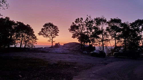 Silhouetted trees against a vibrant sunset sky, with a rocky landscape and parked cars in the foreground.