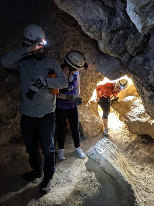 Three people wearing helmets with headlamps explore a rocky cave, with one person examining a narrow opening.