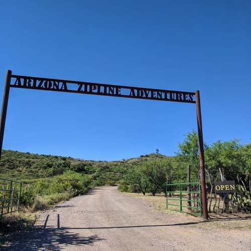 Sign for "Arizona Zipline Adventure" at the entrance of a dirt road surrounded by greenery under a clear blue sky.