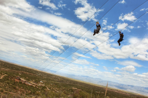 Two people ziplining over a vast landscape with mountains and a cloudy sky in the background.