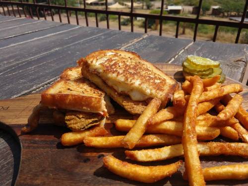 A grilled cheese sandwich with fried chicken, served with crispy fries and a pickle on a wooden platter.