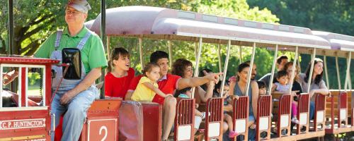 A colorful train with passengers, including children and adults, enjoying a ride through a sunny park.