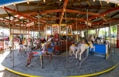 A vintage carousel with colorful wooden horses under a rustic roof, surrounded by bright lights and a festive atmosphere.