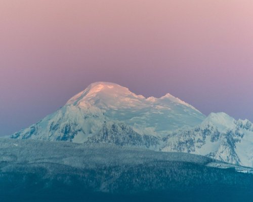 Snow-capped mountain peak bathed in soft pink and purple hues during sunset.