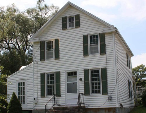 A two-story white house with green shutters and a small porch, surrounded by trees and a clear blue sky.