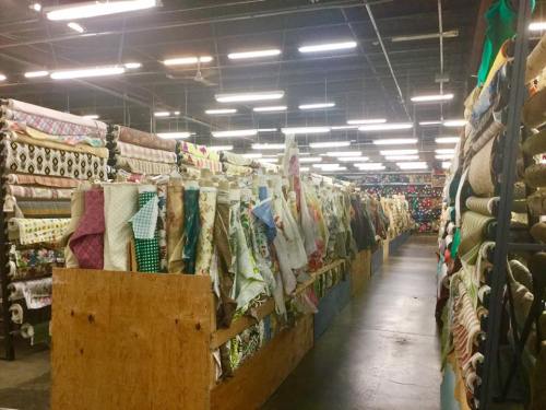 A spacious fabric store aisle lined with colorful rolls of fabric on shelves, illuminated by bright overhead lights.