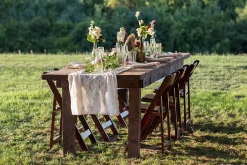 A beautifully set outdoor dining table with flowers, glassware, and a lace runner, surrounded by green grass.