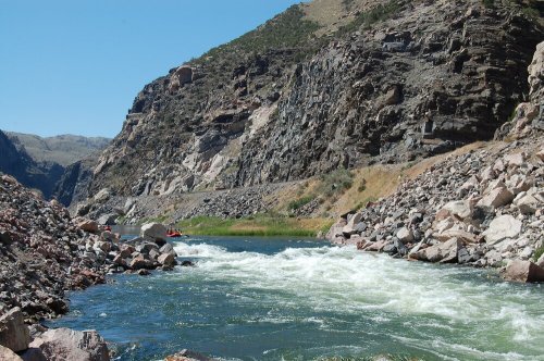 A river flows through rocky cliffs, with a small group of people rafting in the distance under a clear blue sky.