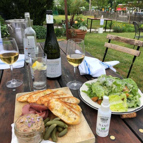 A table set with wine, a salad, charcuterie, and hand sanitizer, surrounded by greenery in a casual outdoor setting.