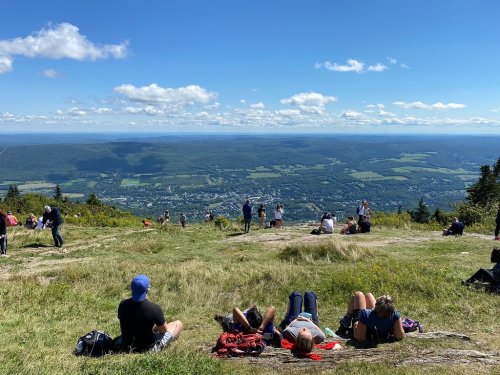 People relax on a grassy mountain slope, enjoying a scenic view of valleys and distant mountains under a clear blue sky.
