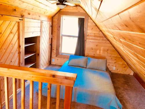 Cozy attic bedroom with wooden walls, a bed covered in a blue blanket, and a window letting in natural light.