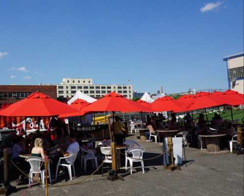 Outdoor dining area with red umbrellas, tables, and chairs, bustling with people enjoying a sunny day.