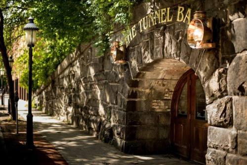 A stone wall with a wooden door, marked "The Tunnel Bar," surrounded by trees and street lamps.